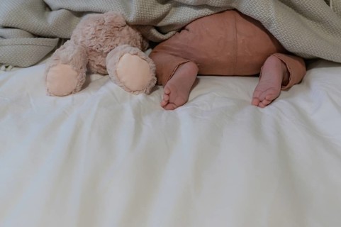 Little baby in brown trousers, next to a plush toy under a blanket on a bed