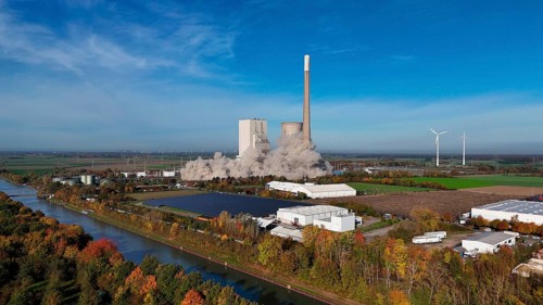 Demolition of a chimney with smoke cloud, surrounded by autumn landscape and wind turbines,