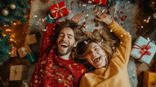 Young couple laying on their backs surrounded by christmas gifts and decorations laughing.