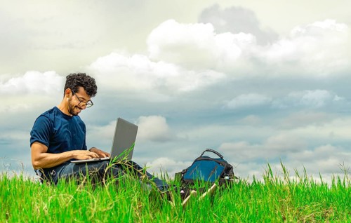 Smiling freelancer man sitting with laptop and backpack in field. Portrait of a man sitting on the