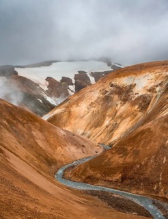 Steaming stream between colourful rhyolite mountains with snowfields, Hveradalir geothermal area,