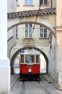 Red historic tram runs through winding houses and alleys with archways in Prague, Czech Republic,