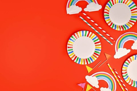 Rainbow themed party flat lay with colorful plates, napkins and drinking straws on red background