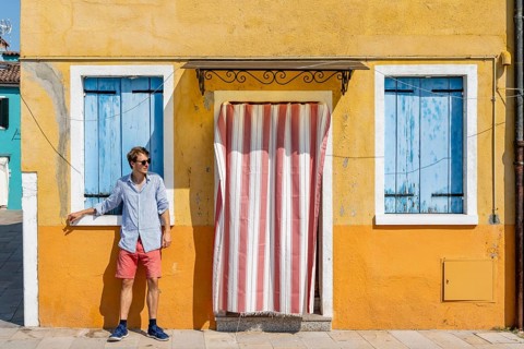 Young man leaning against house wall, yellow house facade with entrance door with red and white