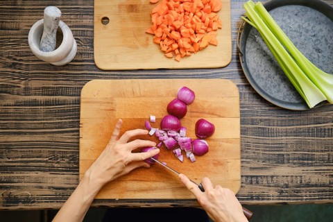 Hands chop red onions on a wooden cutting board with diced carrots and celery nearby. A mortar and