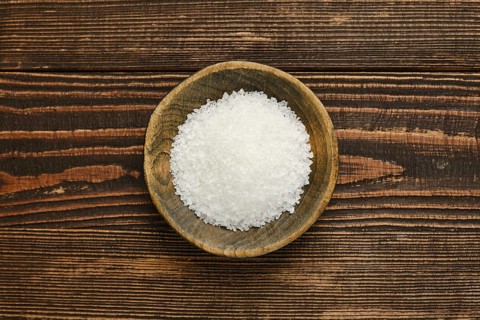Coarse salt is displayed in a rustic wooden bowl, set against a textured wooden table. The natural