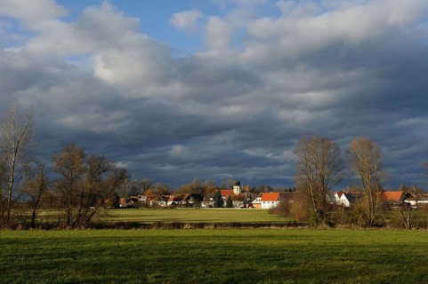 Vast landscape with a small village under a dramatic cloudy sky, surrounded by green meadows and