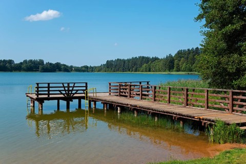 A wooden jetty juts into a calm lake, surrounded by trees and a clear blue sky, Paniewo Lake,