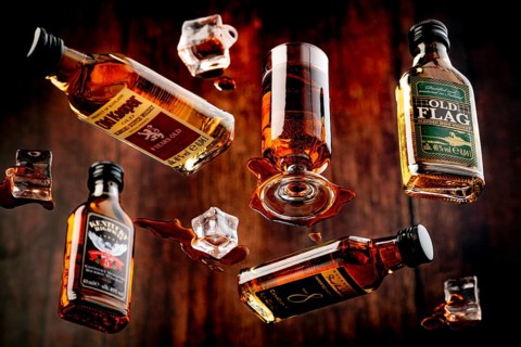 Four whisky bottles and a glass with spilt whisky and melted ice against a wooden background