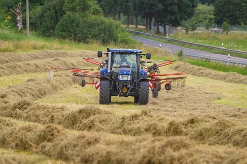 A tractor tilling a hay field next to a country road, surrounded by trees and meadows, Dachtel.