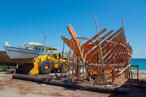 A ship under construction with a yellow excavator at a shipyard by the sea under a blue sky,
