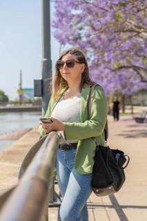 A vertical shot of a woman wearing sunglasses and a green jacket, holding her smartphone with both