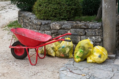 Red metal wheelbarrow next to translucent yellow plastic thrash bags filled with various discarded