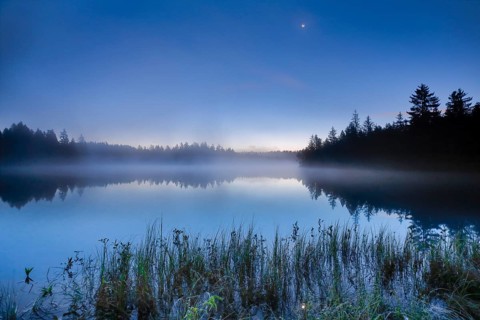 Crescent moon over the mirror-smooth mire lake Étang de la Gruère in the canton of Jura,