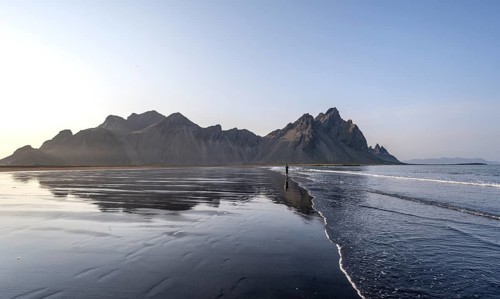 Tourist at black lava beach, sandy beach and sea, mountains Klifatindur reflected in the water,