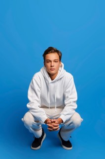 Attractive man wearing white sweatshirt and sweatpants is squatting over blue studio background