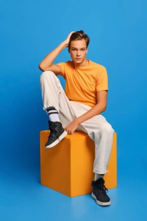 Positive man in orange undershirt and white sweatpants sits on wooden cube over blue studio