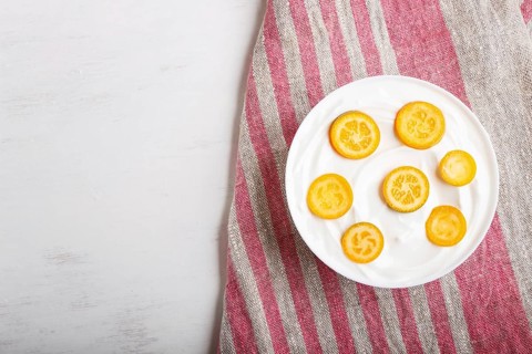 Greek yogurt with kumquat pieces in a white plate on a white wooden background, top view, flat lay,
