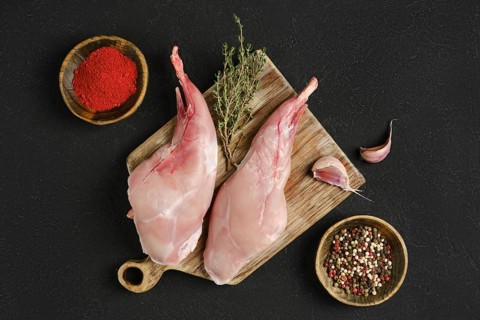 Two whole rabbit legs sit on a wooden cutting board alongside garlic, fresh thyme, and bowls of