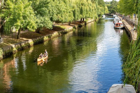 Boats moored people kayaking, River Wensum, Norwich, Norfolk, England, UK treelined riverbank