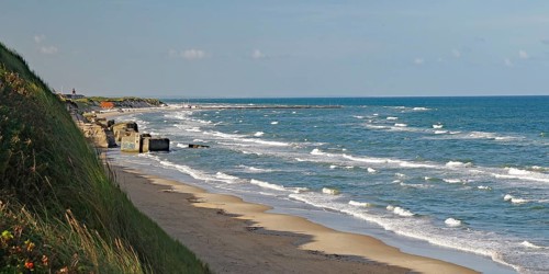 View of the beach with gentle waves lapping against the shore, overgrown with grass and sand dunes,