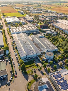 An industrial area with several buildings, roads and solar modules, surrounded by fields, taken
