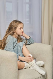 A young pensive girl sits in profile on a soft armchair in a well-lit room with her hand resting