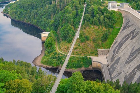 View of a bridge crossing a dam surrounded by dense forest and a calm body of water,
