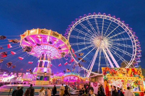 A lively funfair at dusk, illuminated by a large Ferris wheel and a colourful chain carousel,