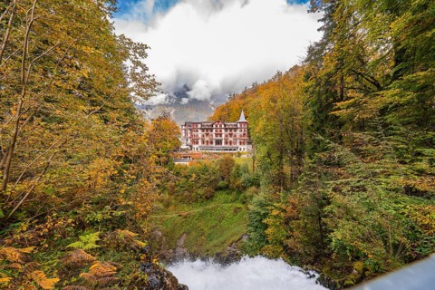 A large hotel surrounded by autumnal trees with mountains and clouds in the background, Lake