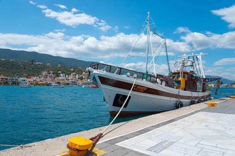 A fishing boat moored on a sunny pier overlooking picturesque hills and coastal houses, Poros,