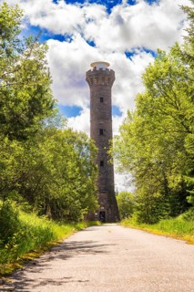 A tall stone tower rises above a road, flanked by trees under a sky full of clouds and sunbeams,