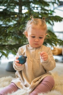 Toddler in a beige dress holding a Christmas tree bauble in front of a festively decorated tree