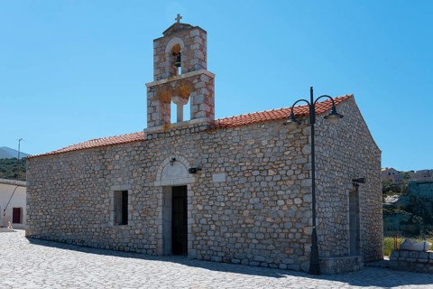 Historic stone church with bell tower and red roof under a blue sky in a Mediterranean setting,