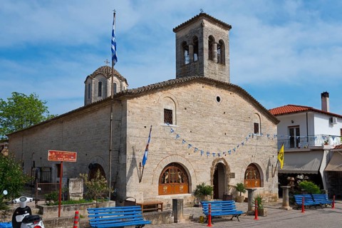 Historic church with bell towers and Greek flags, surrounded by blue benches under a blue sky,