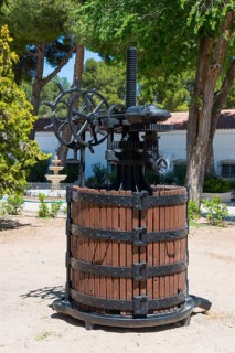 Historische Weinpresse aus Holz und Metall in einem sonnigen Garten mit Bäumen und einem Brunnen,