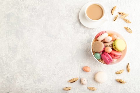 Multicolored macaroons and chocolate eggs in ceramic bowl, cup of coffee on gray concrete