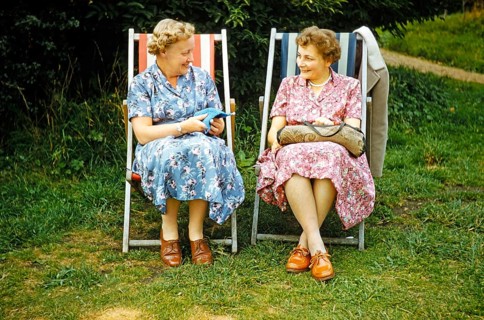 Two women friends sitting in deckchairs on graß lawn one knitting, smiling at each other, England,
