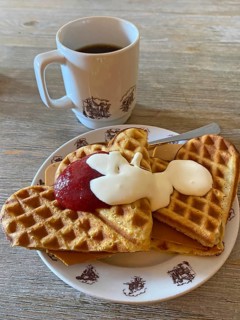Waffles with jam, cream and cheese on a plate, next to a cup of coffee, served on a wooden table,