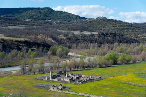 Ruins of an old building in a green field surrounded by a river and wooded hills on a clear spring