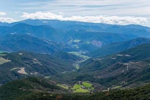 Weitreichende Berglandschaft mit einem grünen Tal, bewaldeten Hängen und einem blauen Himmel mit