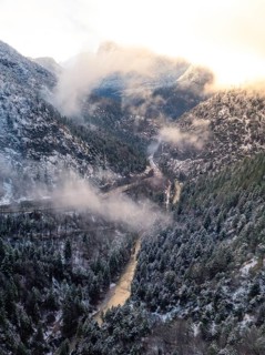 A valley with a river flowing through it surrounded by snow-covered mountains and wafts of mist,