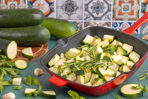 Diced courgettes in a red pan, surrounded by fresh herbs and whole courgettes in front of colourful