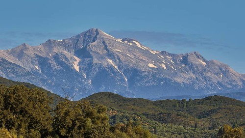 A majestic, snow-capped mountain peak rises above green forests, Taygetos Mountains, Mani