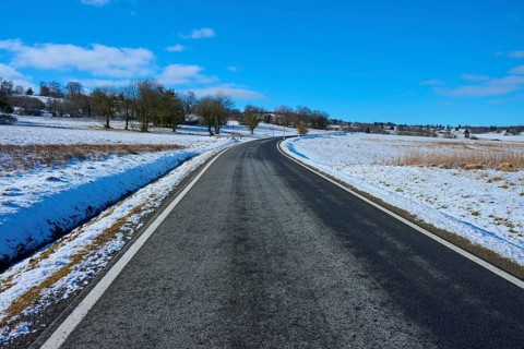 An empty country road leads through a wintry landscape with snow-covered trees and blue sky,
