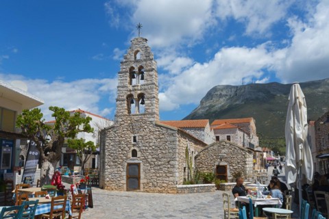 Small church with bell tower in a lively street with cafés and mountains in the background, double