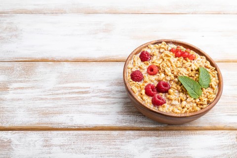 Wheat flakes porridge with milk, raspberry and currant in wooden bowl on white wooden background.