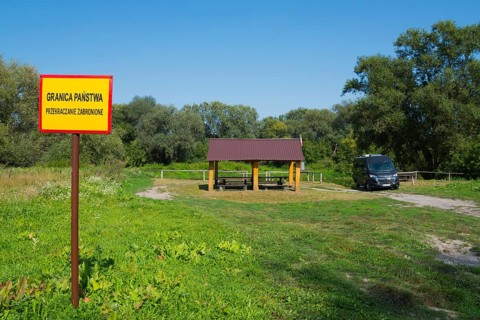 Border sign next to a wooden shelter on a green field with trees and car, motorhome, camper, border