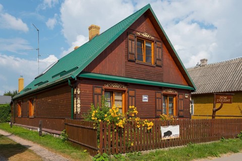 Dark brown wooden house with green roof and flowers in the garden behind the fence, Bialowieza,