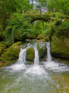 An idyllic waterfall flows under a rustic bridge, surrounded by lush greenery and rocks, aerial